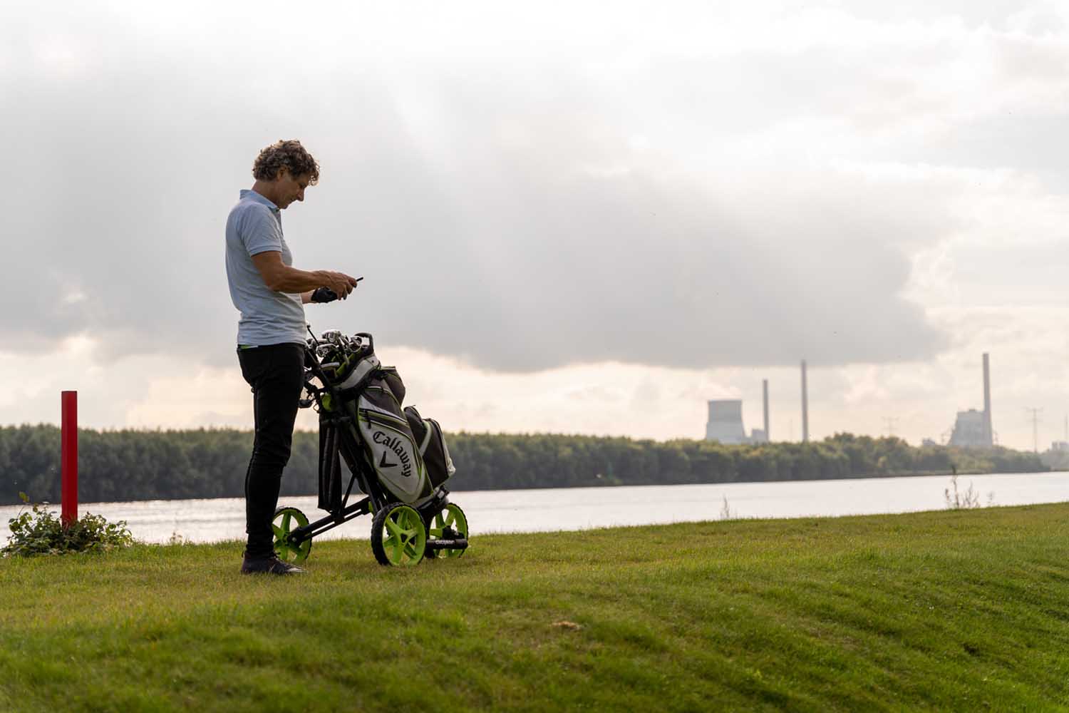 Foto van man op dijk met golfclubs en mobiel in hand, op de achtergrond Amercentrale Geertruidenberg